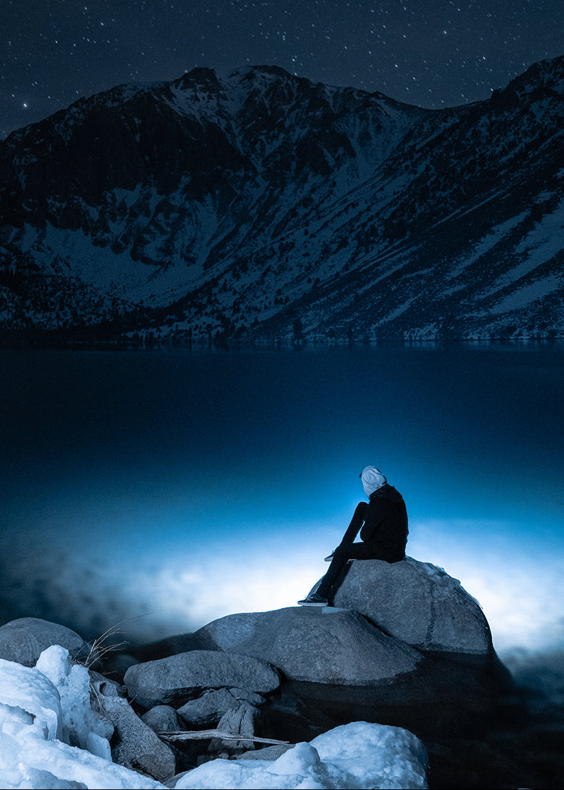 A person in dark clothing sits on a large rock at the edge of a frozen lake under a starry night sky, with snow-covered mountains in the background. The scene is dominated by deep blue and black tones with bright icy highlights.