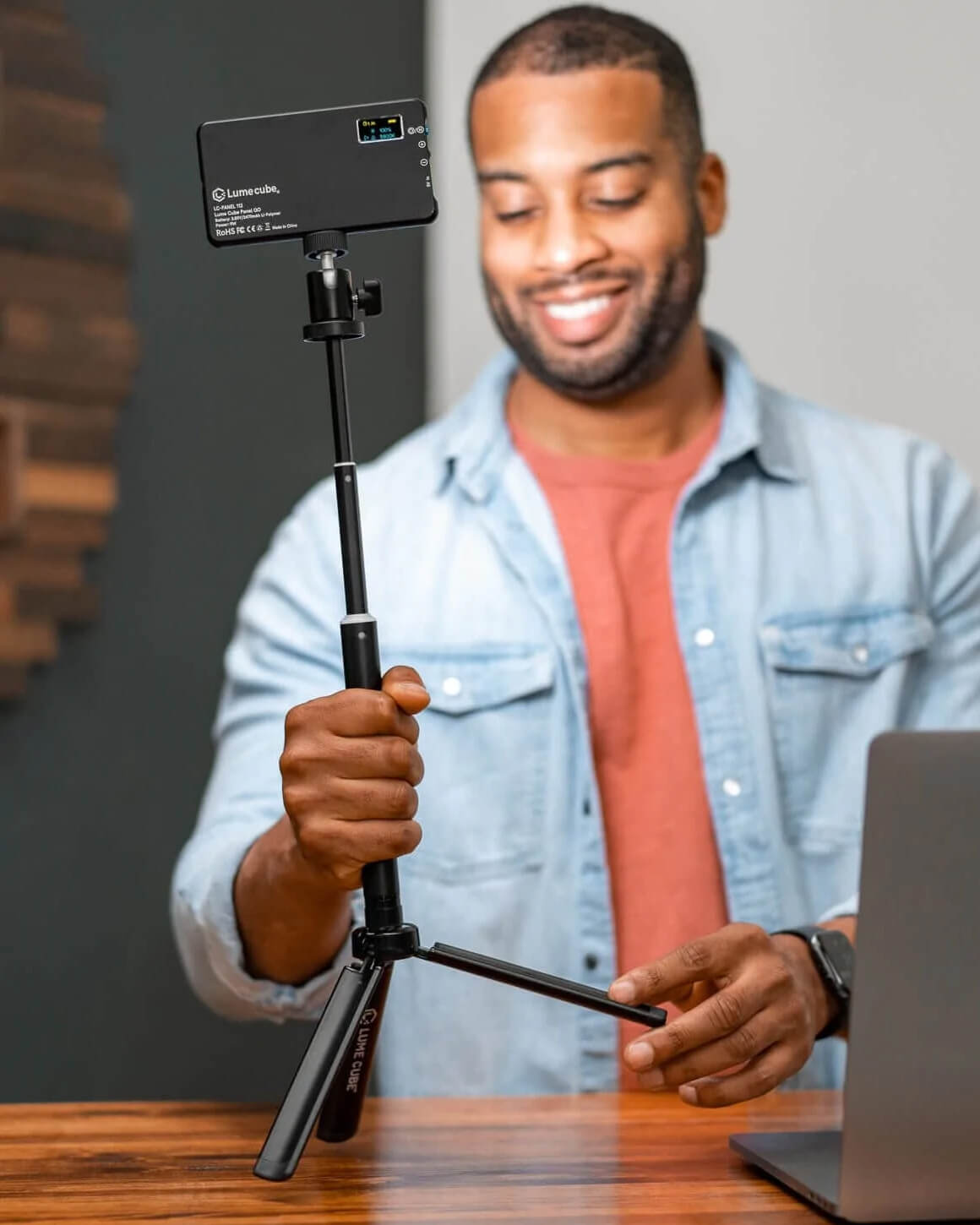 Man setting up Lume Cube's 30" black metal Adjustable Light & Webcam Stand with Rotating Mount next to his laptop while fully-extended with a LED panel attached. LC-STAND
