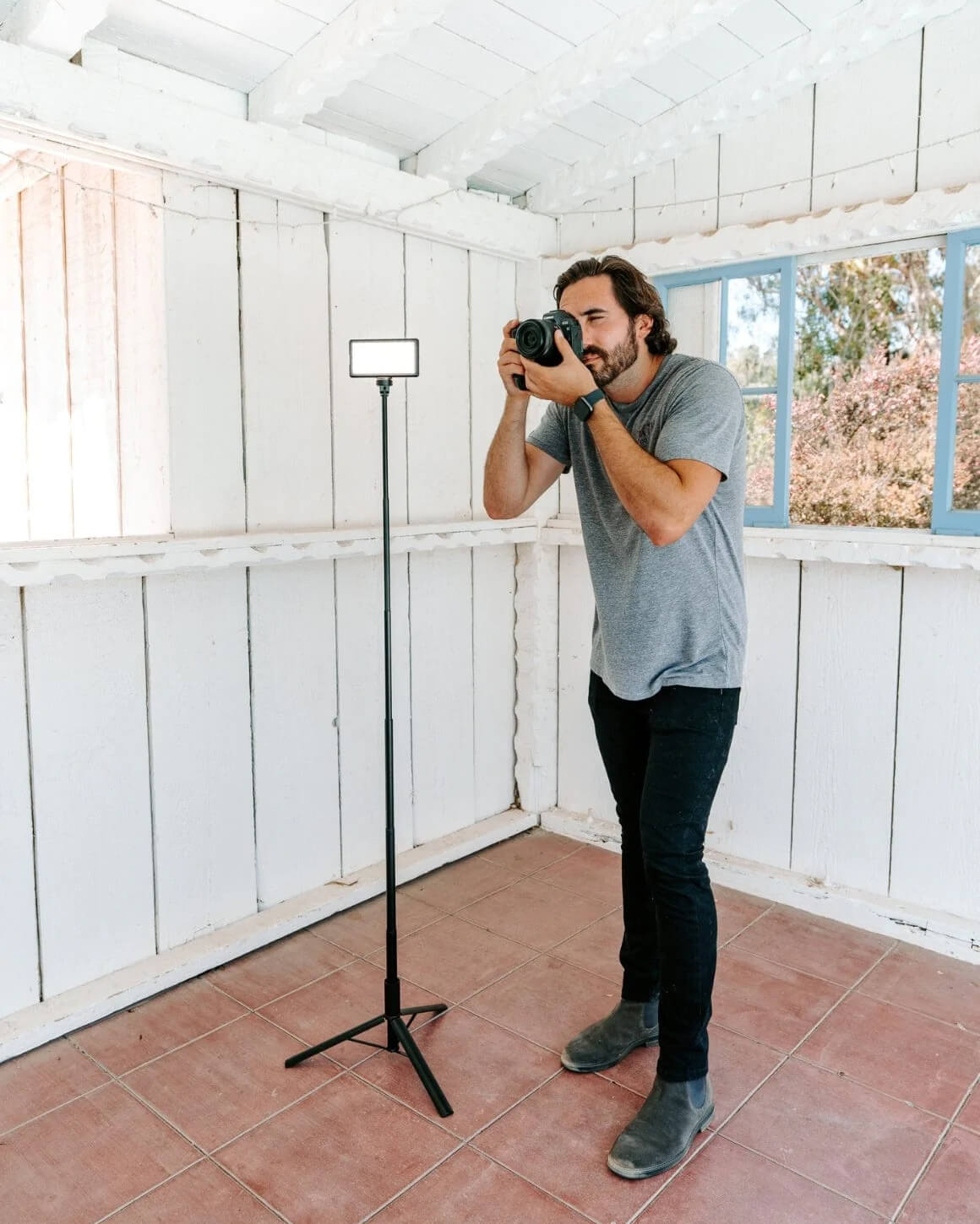 Man standing to take photos aside black metal Lume Cube 5-Foot Adjustable Light Stand with an RGB Panel Pro attached. LC-STANDT60