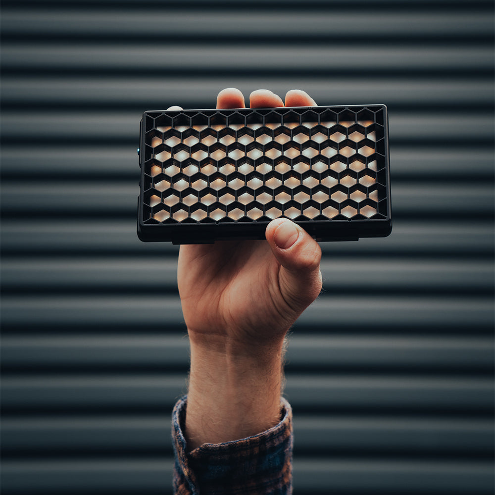 A hand holding a rectangular black honeycomb-patterned makeup compact against a dark, horizontally ribbed background.