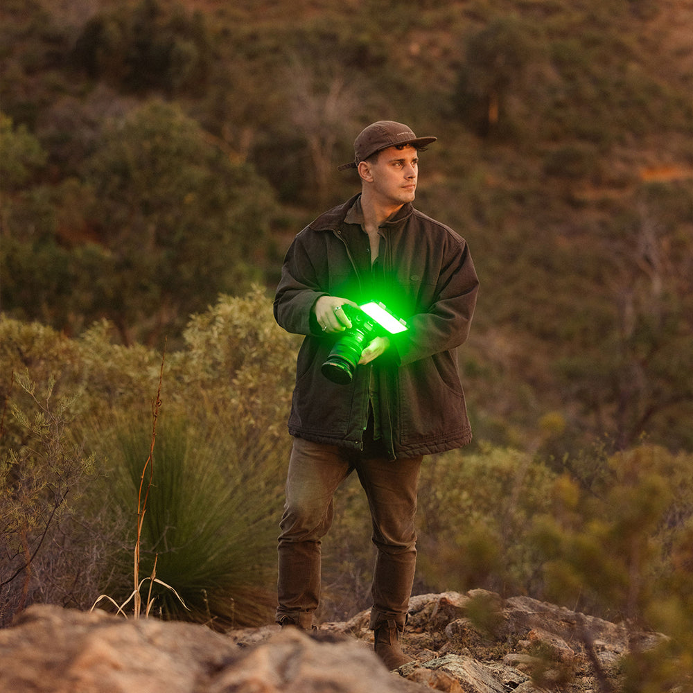 A man standing outdoors in a rocky, brush-filled landscape at sunset, holding a green lighted camera or flashlight. He is wearing a dark jacket, brown pants, and a cap.