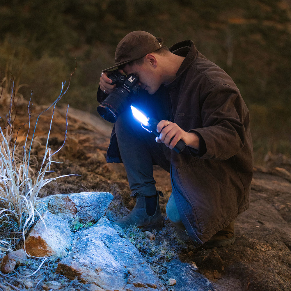 A man crouches outdoors at night holding a camera and a bright blue LED flashlight, illuminating rocks and dry grass in a natural setting.
