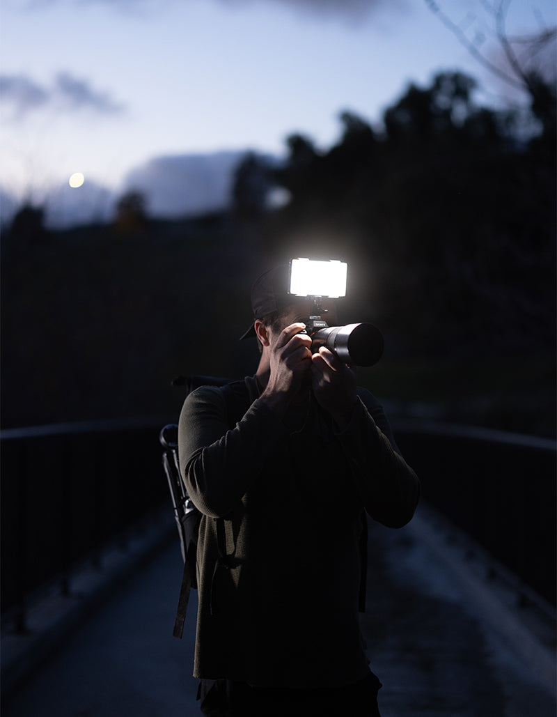 A person standing on a bridge at dusk, holding a camera with a bright rectangular light attached to the lens, against a dark outdoor background.