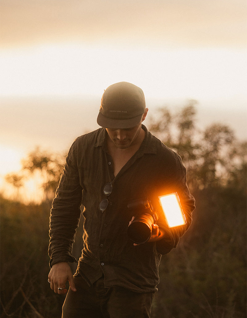 A man wearing a dark cap and button-up shirt stands outdoors at sunset holding a camera with a bright rectangular light attached, against a warm golden sky.