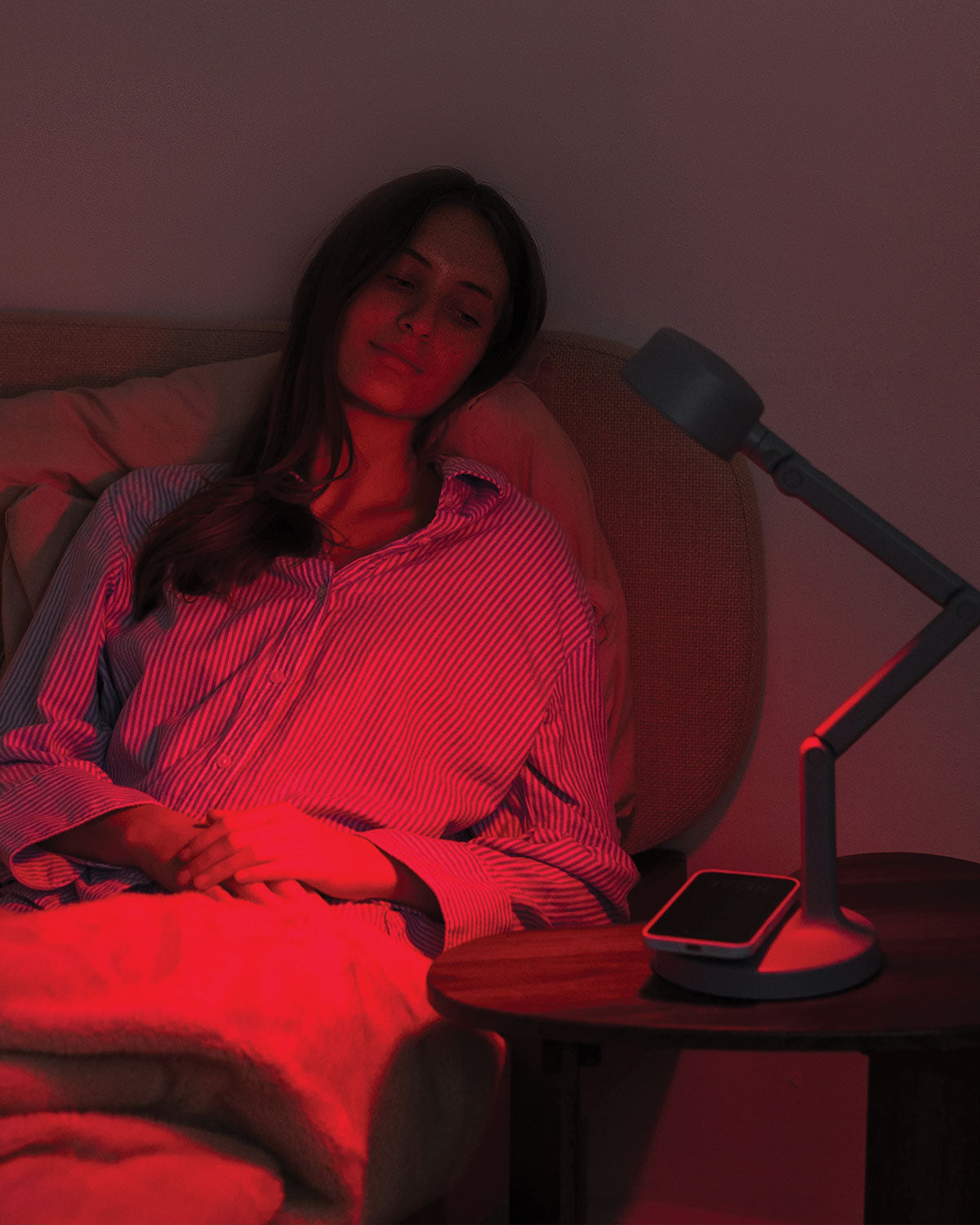 Woman lying in bed under red lighting with a portable desk lamp and phone on the nightstand.