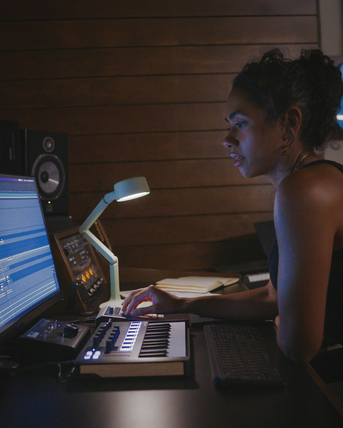 Person using a music production setup with a computer and keyboard in a dimly lit room in light of a rechargeable desk lamp